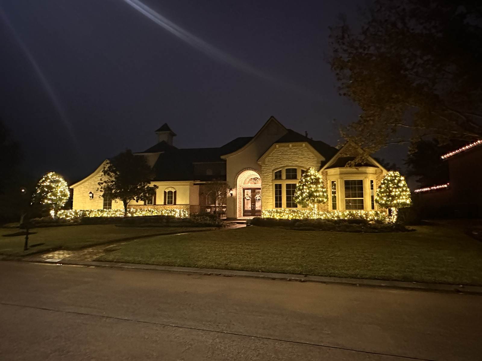Warm white lighting along a landscaped estate home's roofline and trees at dusk